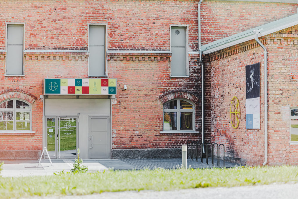 Taide- ja Kulttuurikeskus Kalevan Navetan pääsisäänkäynti Itikanmäen sisäpihalta päin kuvattuna. Pääoven oikealla puolella on pyörätelineitä ja oven päällä pieni koristeellinen lippa.