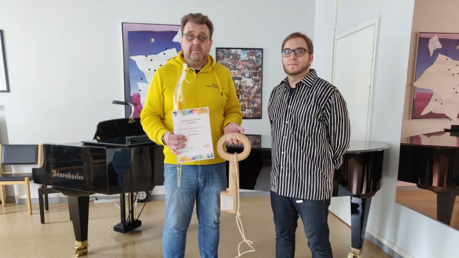 kaksi miestä seisovat, joista toinen pitelee avaimen muotoista palkintoa