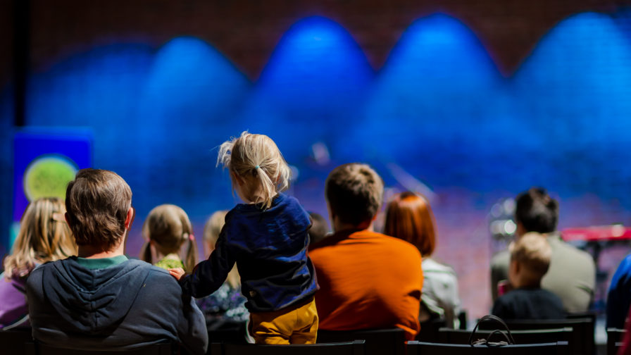 Lapsiperheistä koostuvaa yleisöä selkäpuolelta kuvattuna. Taustalla näkyy Hugo-salin tiiliseinää. jossa sinisiä valokeiloja.