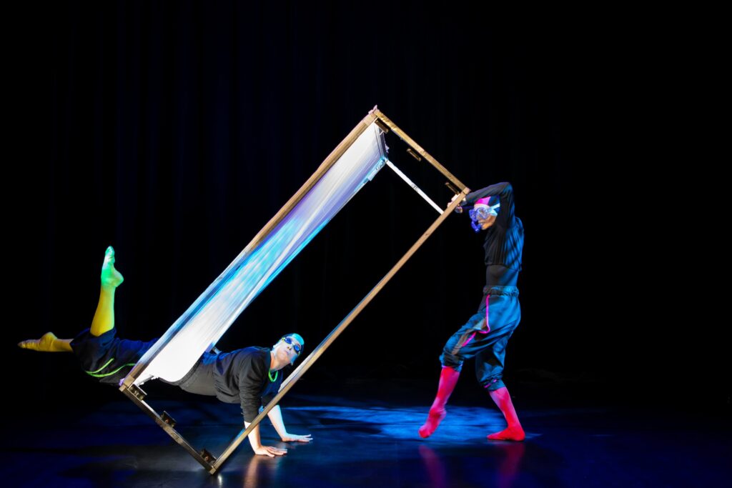 Kaksi ihmistä suorakaiteen muotoisen kehikon ympärillä. Toinen pitää laatikosta kiinni tukien sitä, ja toinen roikkuu kehikon reunalta jalat ilmassa liikkuen.